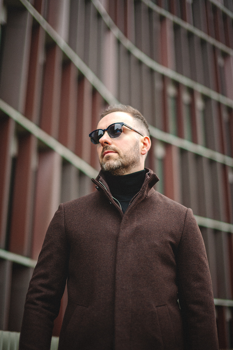 A man in sunglasses and a brown coat against the backdrop of the Panum building in Copenhagen