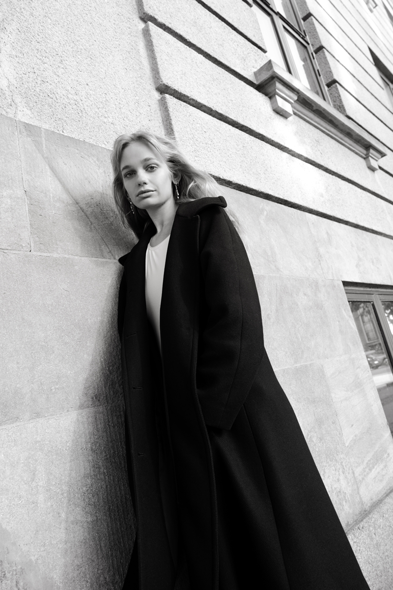 A woman in a black coat leaning sideways against a townhouse wall