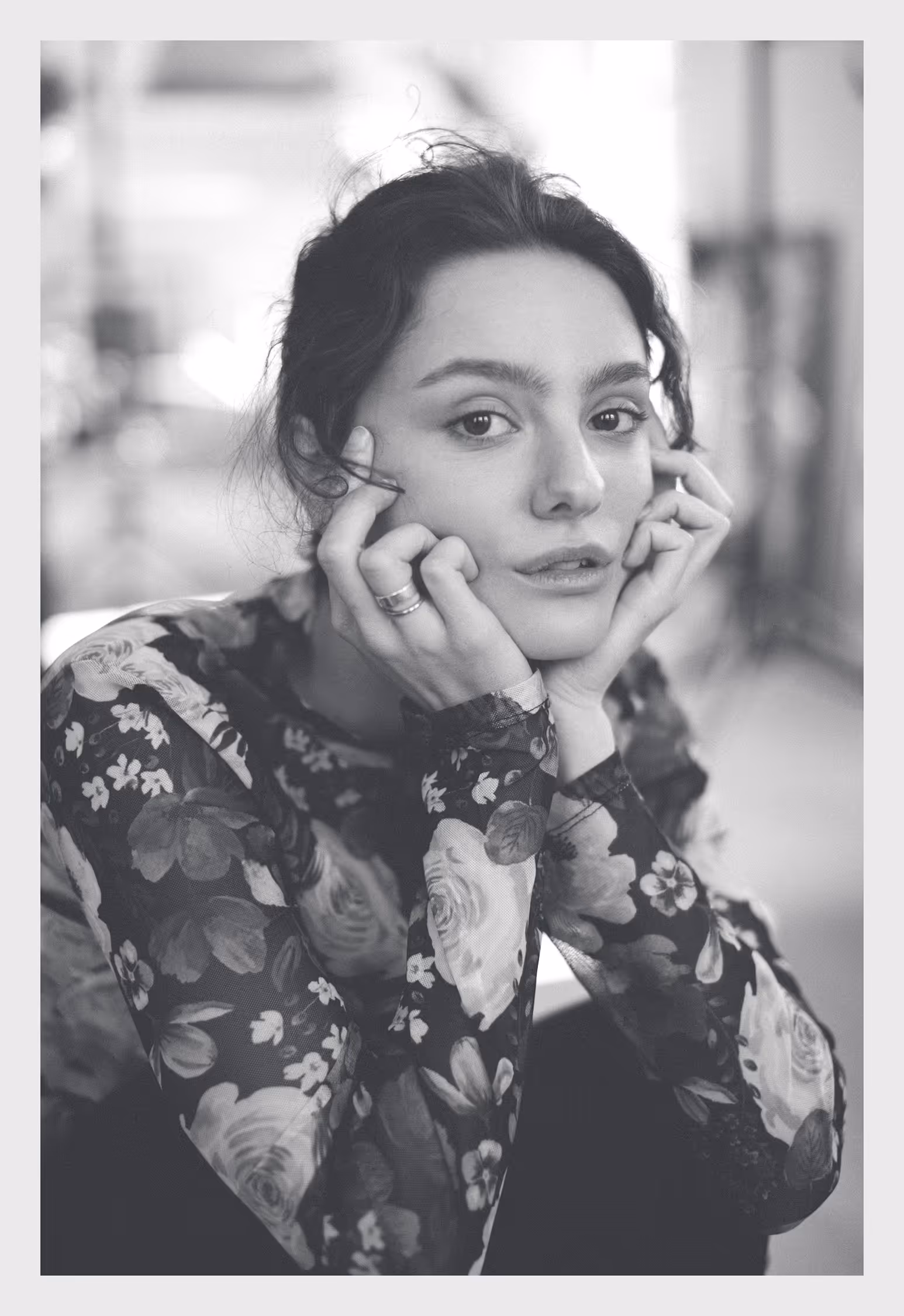 A young woman rests her face in her hands, wearing a floral blouse, looking softly at the camera