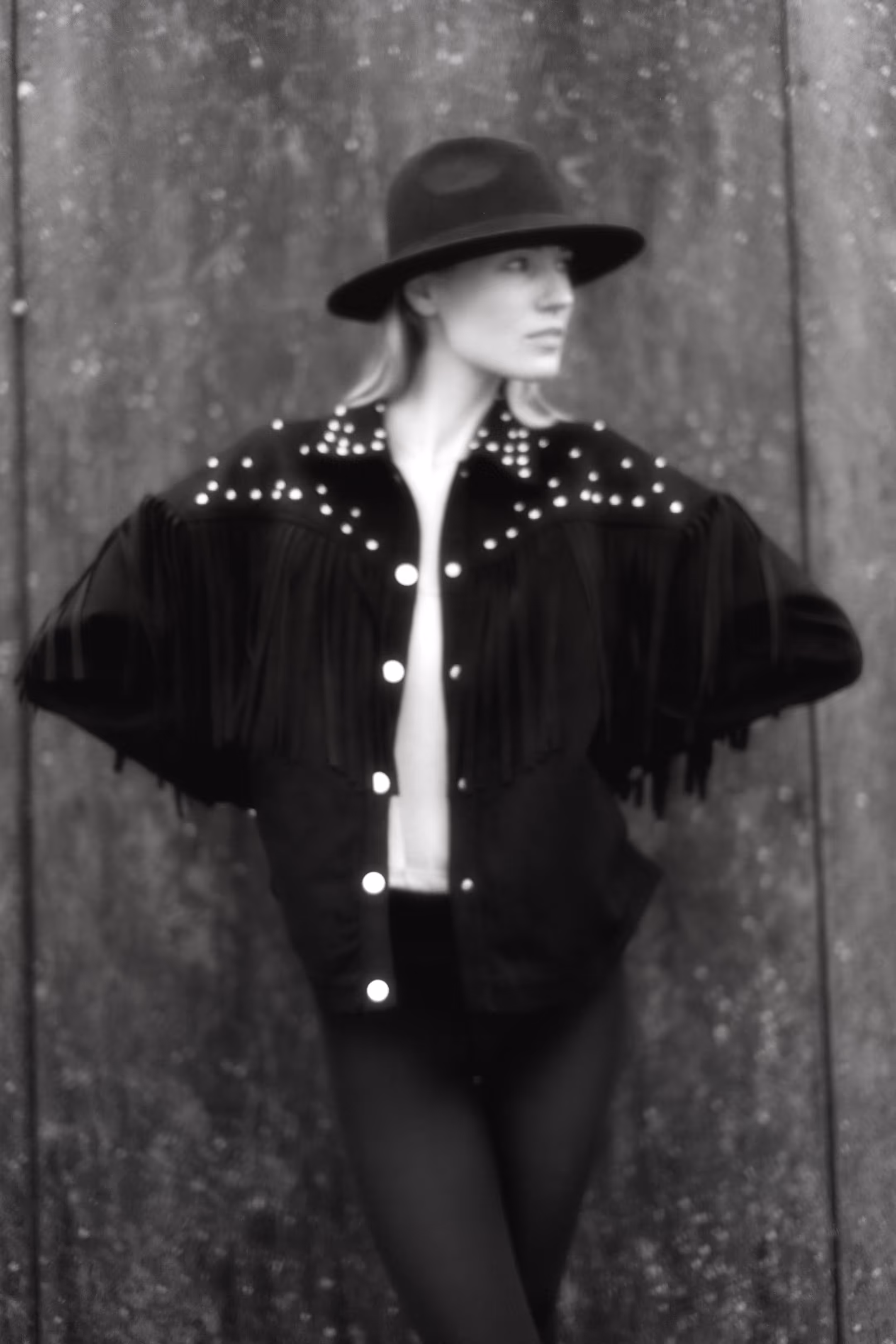 A woman in a hat and fringed jacket, looking to the side, standing against a wall.