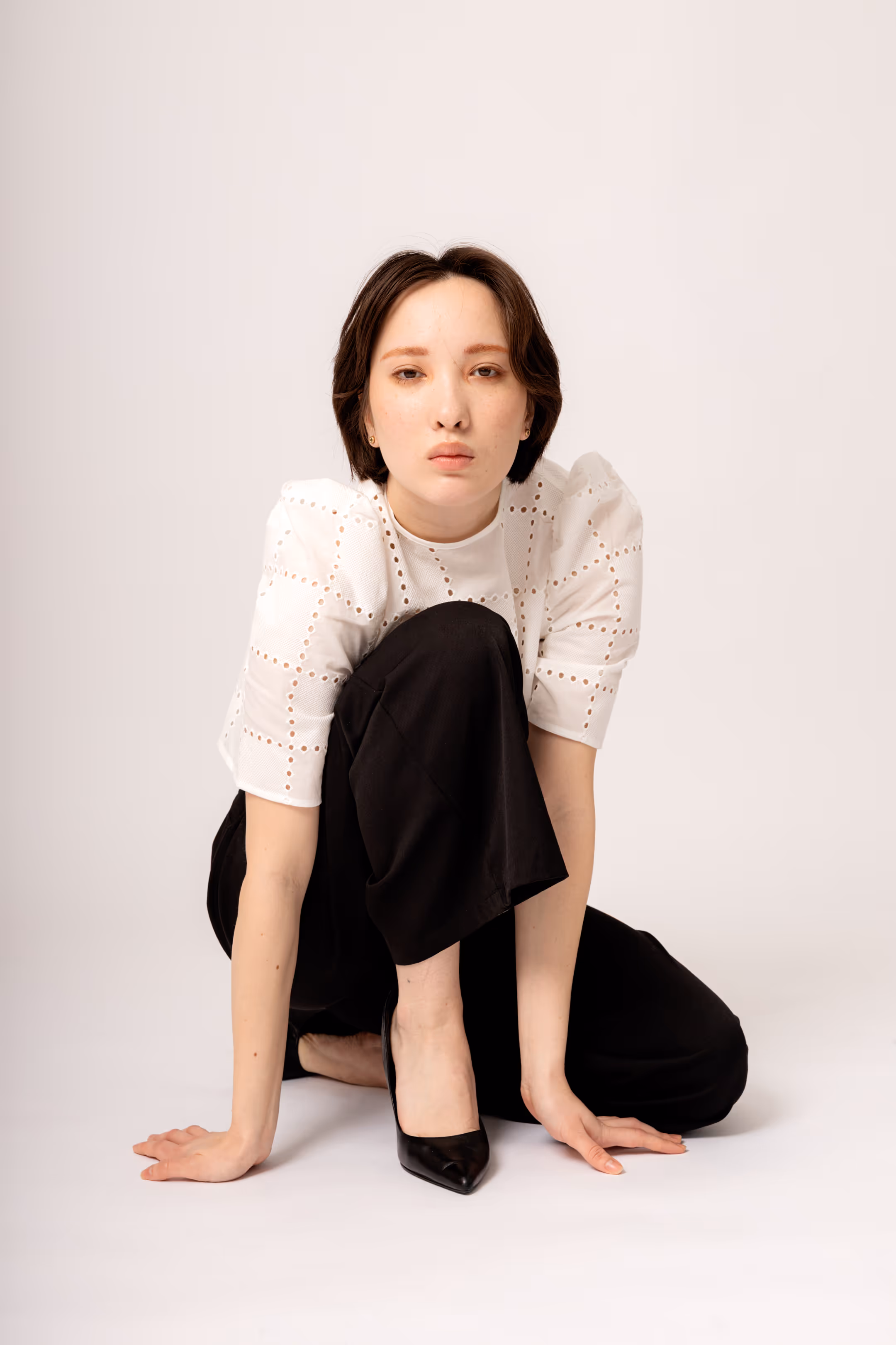 A woman in a white blouse and black pants, sitting on a white photography backdrop, supporting herself with her hands