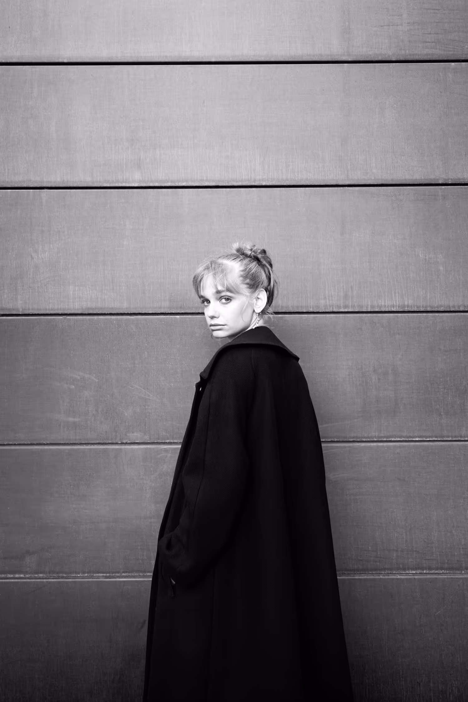 Black and white minimalist portrait in Aarhus. A model wears an oversized black coat, creating a strong silhouette against a linear architectural background with horizontal paneling.
