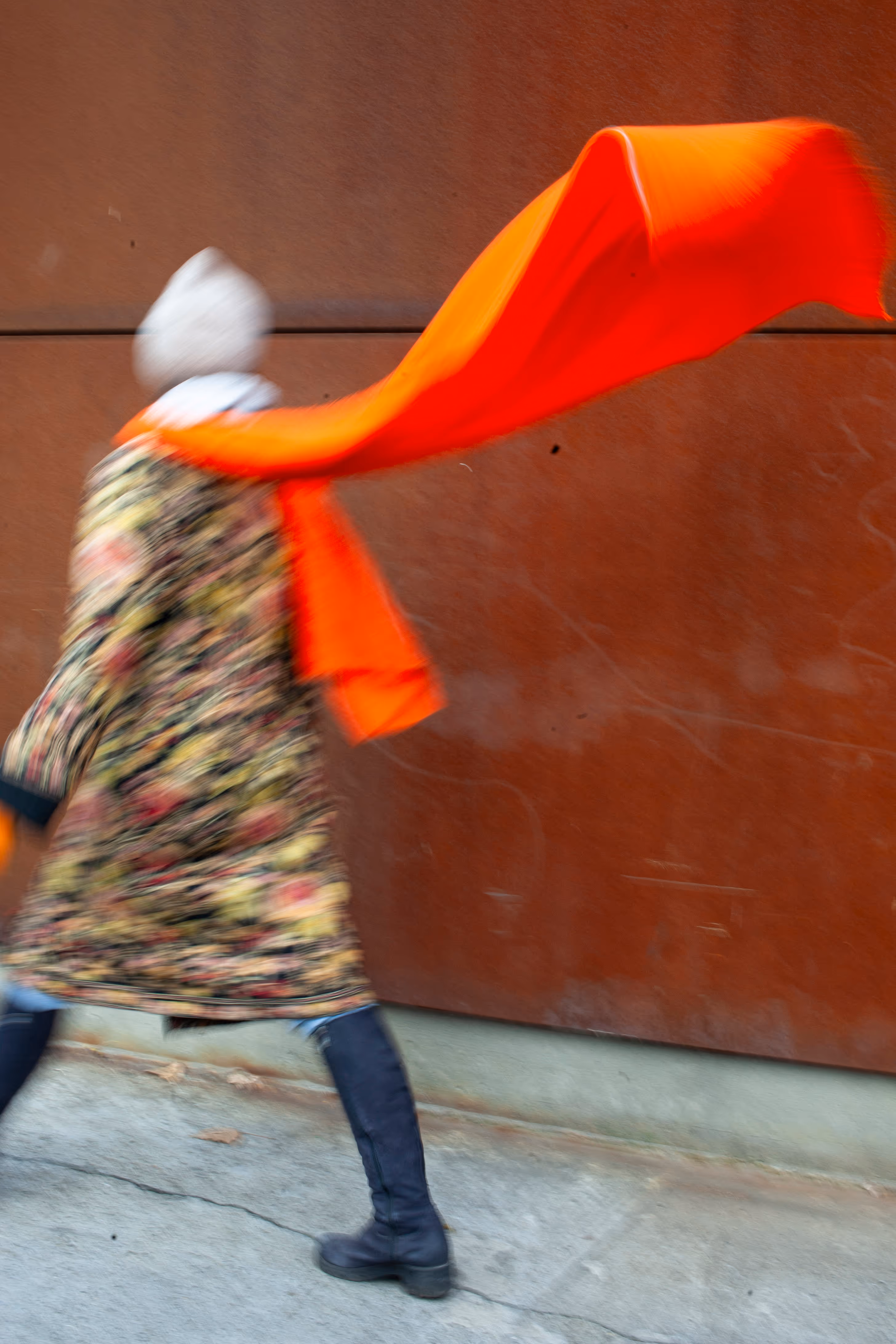 Abstract street photography featuring a person walking with a long, flowing bright orange scarf. The image uses intentional motion blur to create a dynamic, painterly effect.