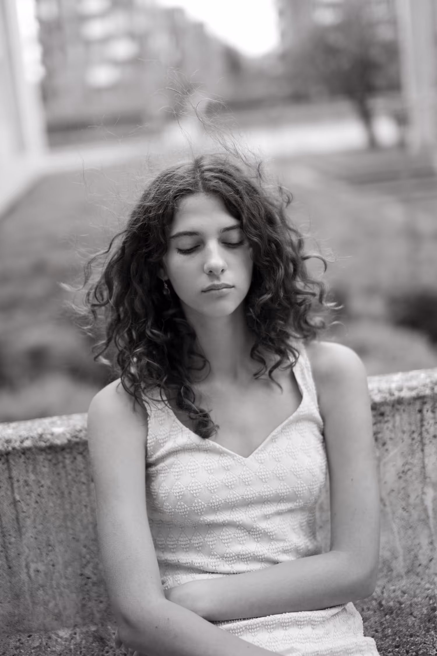 Black and white atmospheric portrait taken at Rødkilde Gymnasium in Vejle. A young woman with curly wind-blown hair stands with closed eyes, creating a serene, ethereal moment with soft bokeh background.