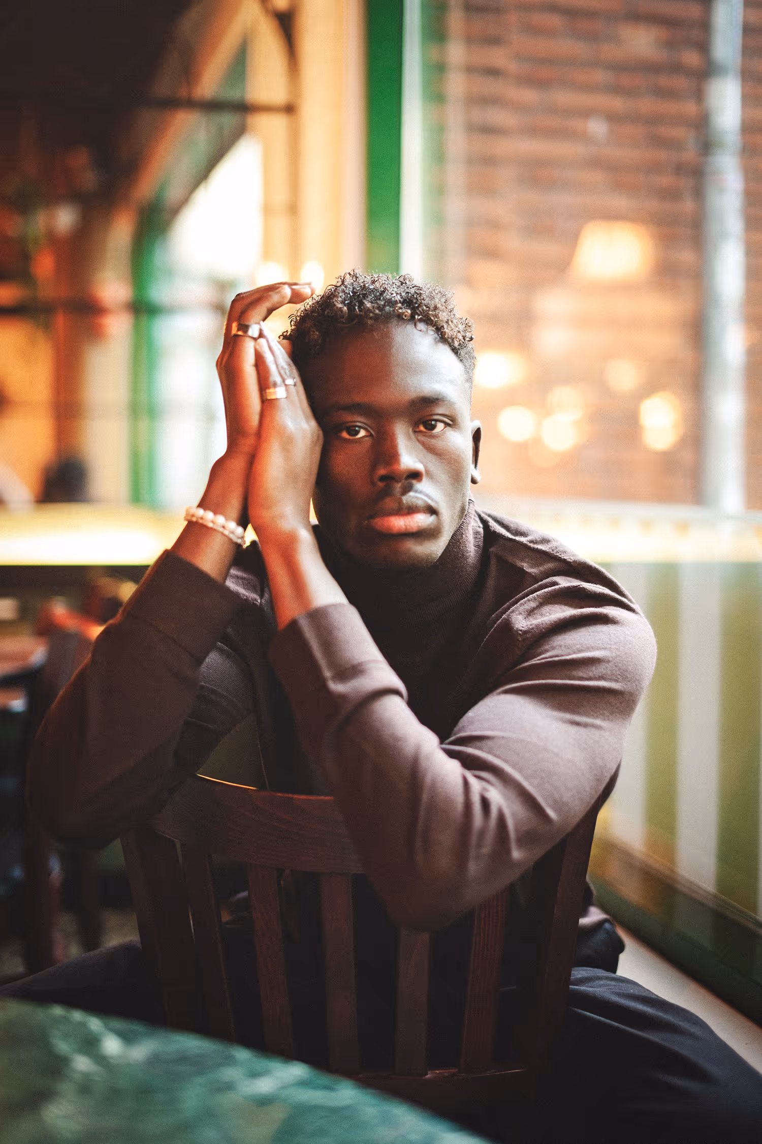 Intimate, cinematic fashion portrait of a man wearing a brown turtleneck and jewelry, sitting at a table in Cafe Krone, Aarhus.
