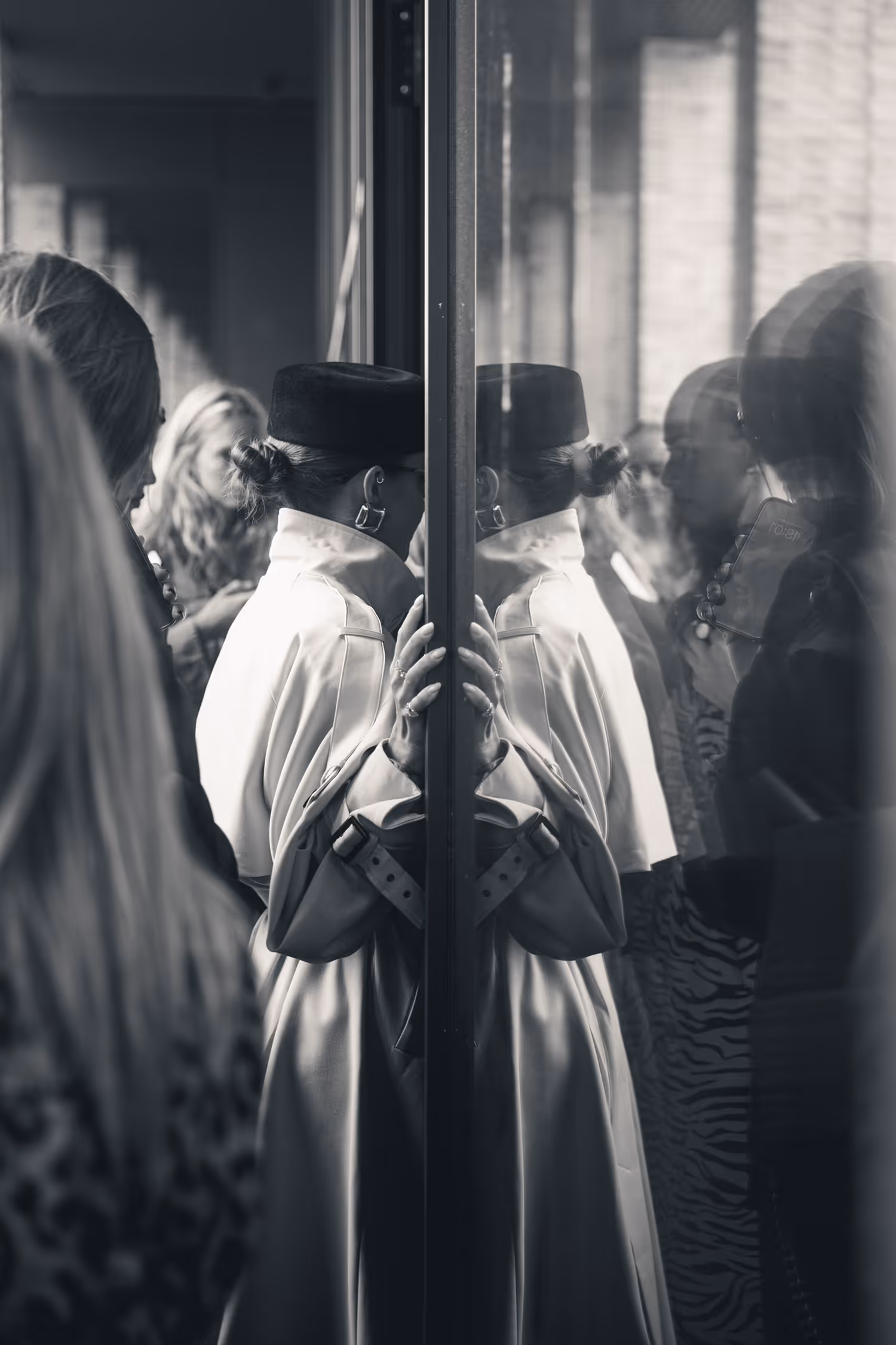 Monochromatic street style photograph captured backstage at Copenhagen Fashion Week 2025. It shows a woman in a trench coat reflected in glass, utilizing high contrast and hard light to emphasize the moment and surrounding fashion crowd.