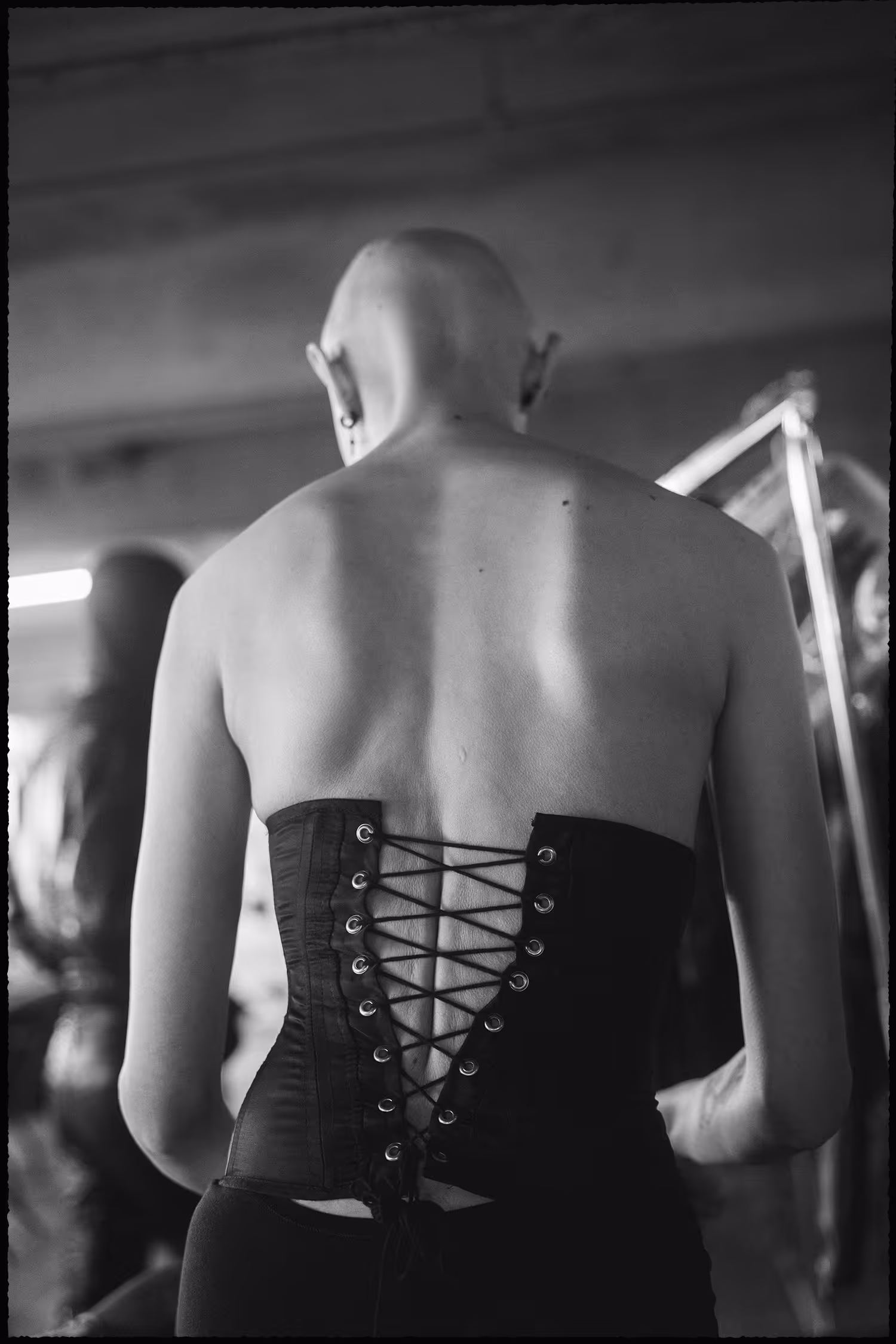 Documentary B&W backstage shot at Han Kjøbenhavn, Copenhagen Fashion Week 2025. A bald model is captured from behind wearing a structured laced corset in a raw, grainy atmosphere.