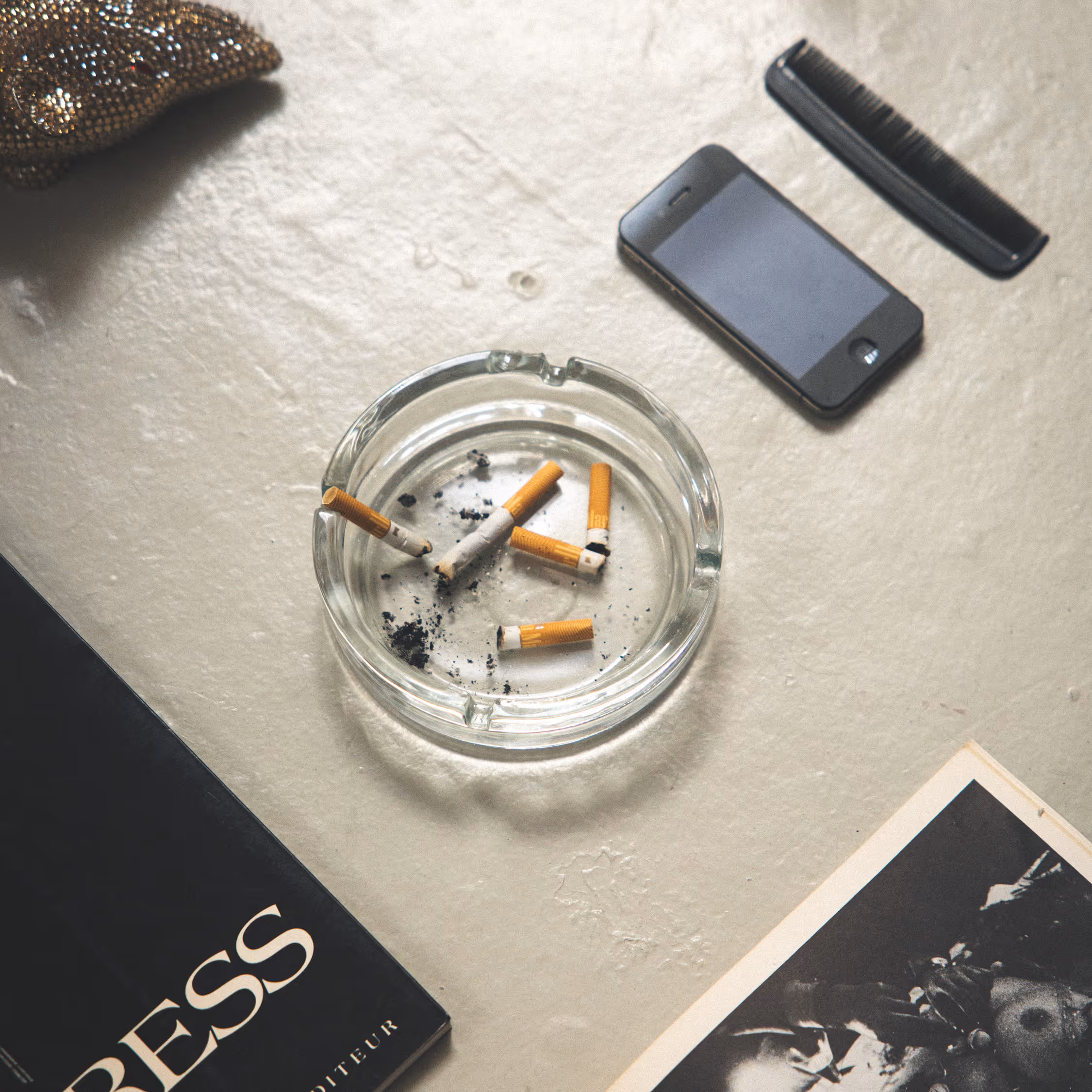 Raw backstage still life from Copenhagen Fashion Week 2025. Top-down view of a glass ashtray with cigarettes, a smartphone, a comb, and "PRESS" materials on a textured surface.