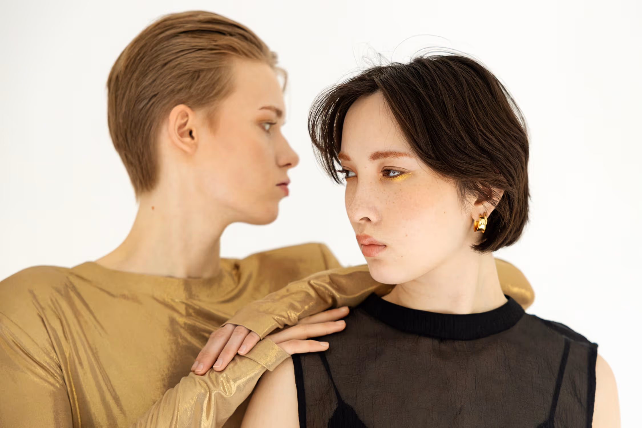 High-key, contrasting fashion portrait of two models in Vejle; one wears metallic gold fabric, the other sheer black mesh, with soft, directional studio lighting.