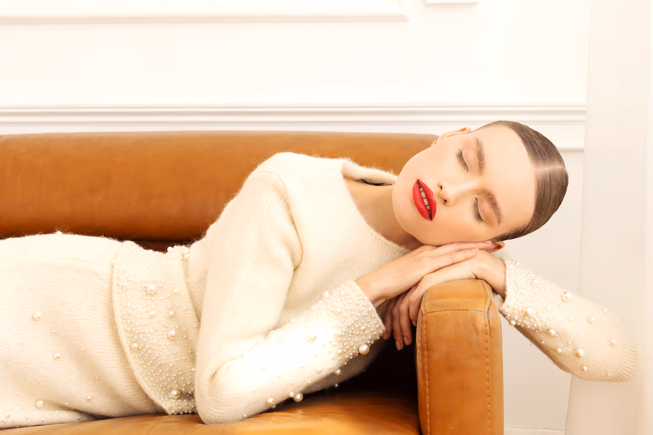 High-key fashion editorial portrait in Warsaw; a model in pearl-embellished knitwear is resting on a cognac leather sofa, highlighted by bright studio light and bold red lips.