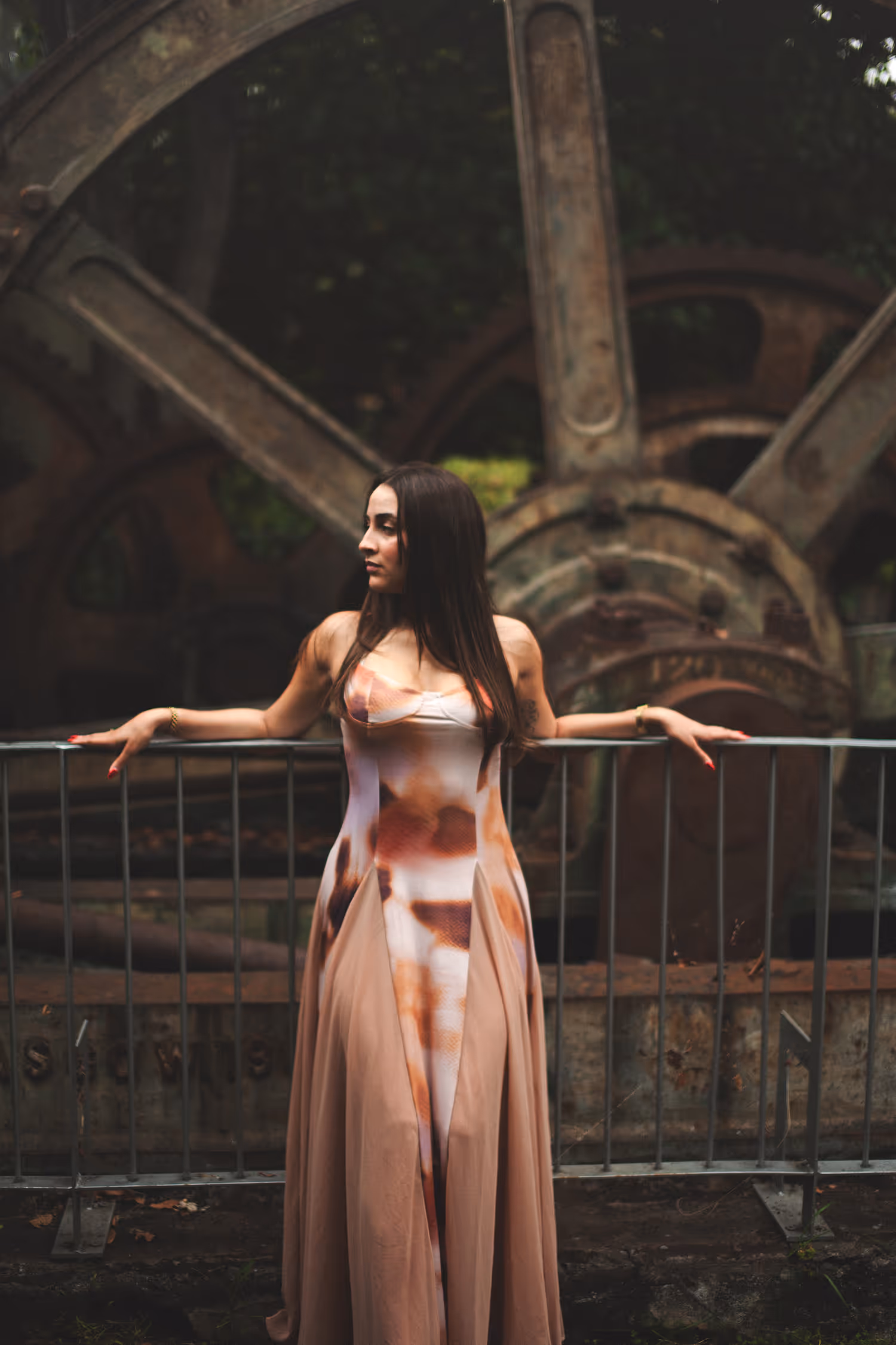 Atmospheric fashion portrait in Funchal's Santa Luzia Garden. A model in an earth-tone dress poses against massive rusted industrial gears in soft, diffused light.