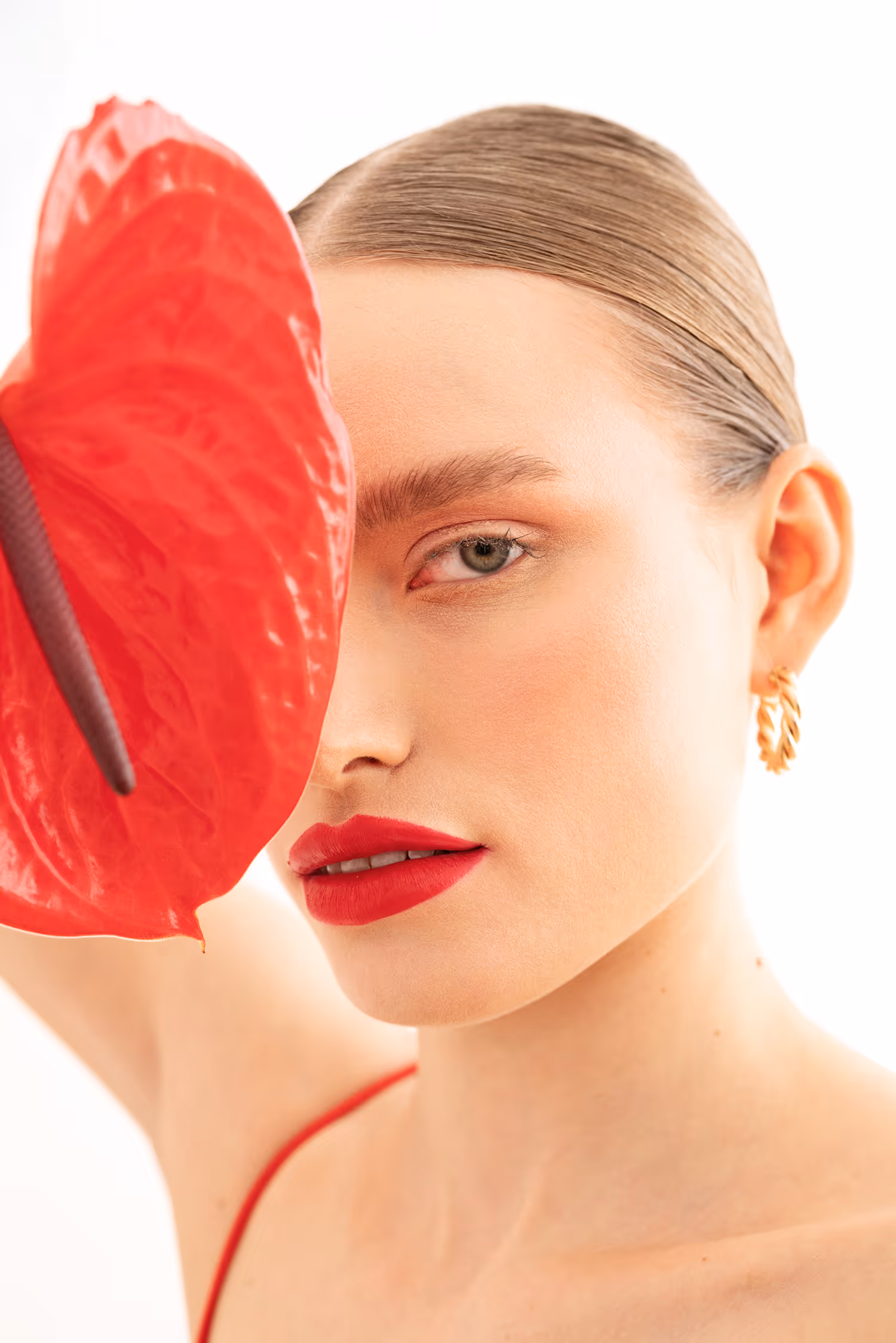 High-key beauty portrait in Warsaw featuring a model with bold red lips covering one eye with a vibrant red anthurium flower.