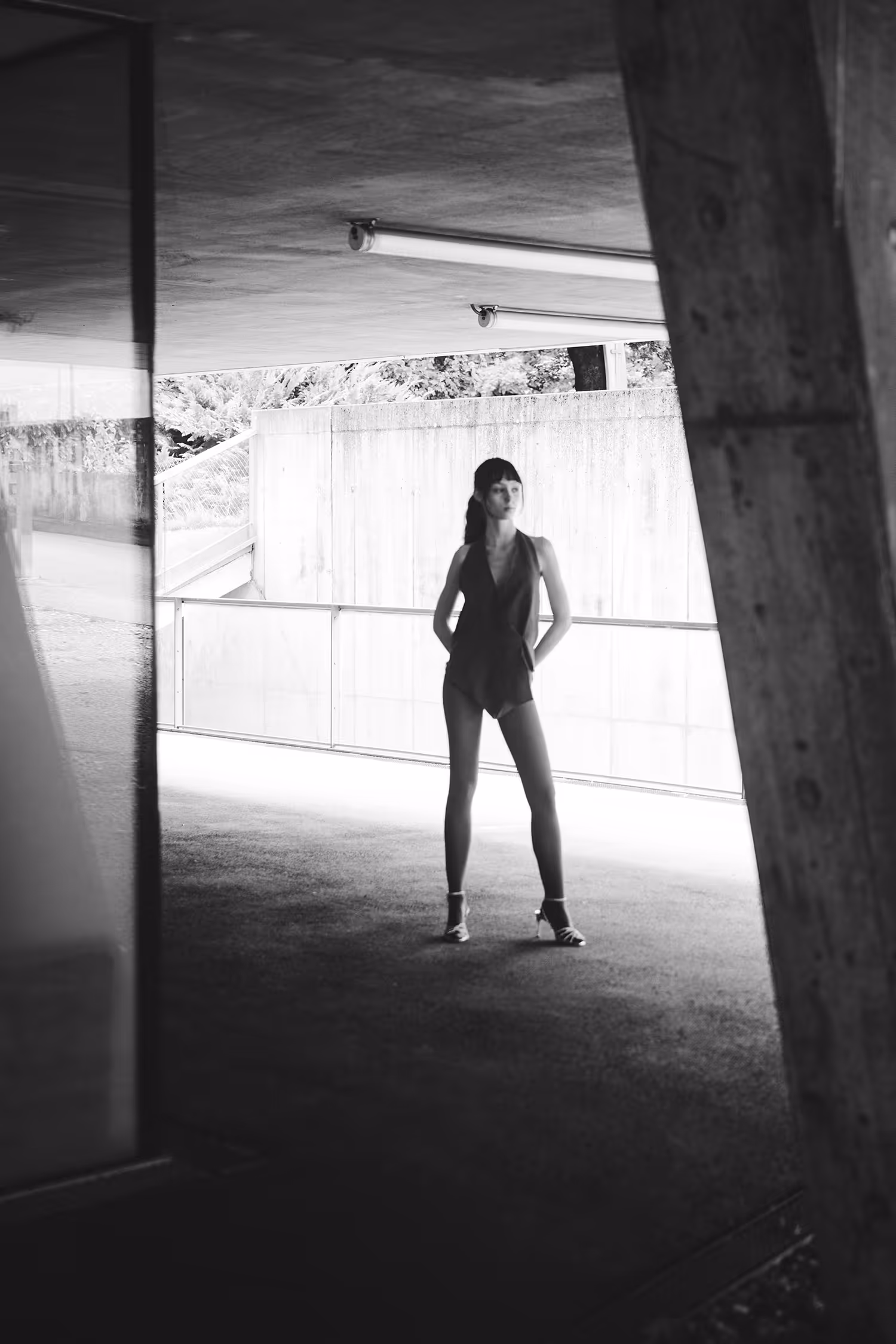 Full-body B&W fashion shot in Innsbruck, featuring a model framed by raw brutalist concrete architecture and natural backlighting.