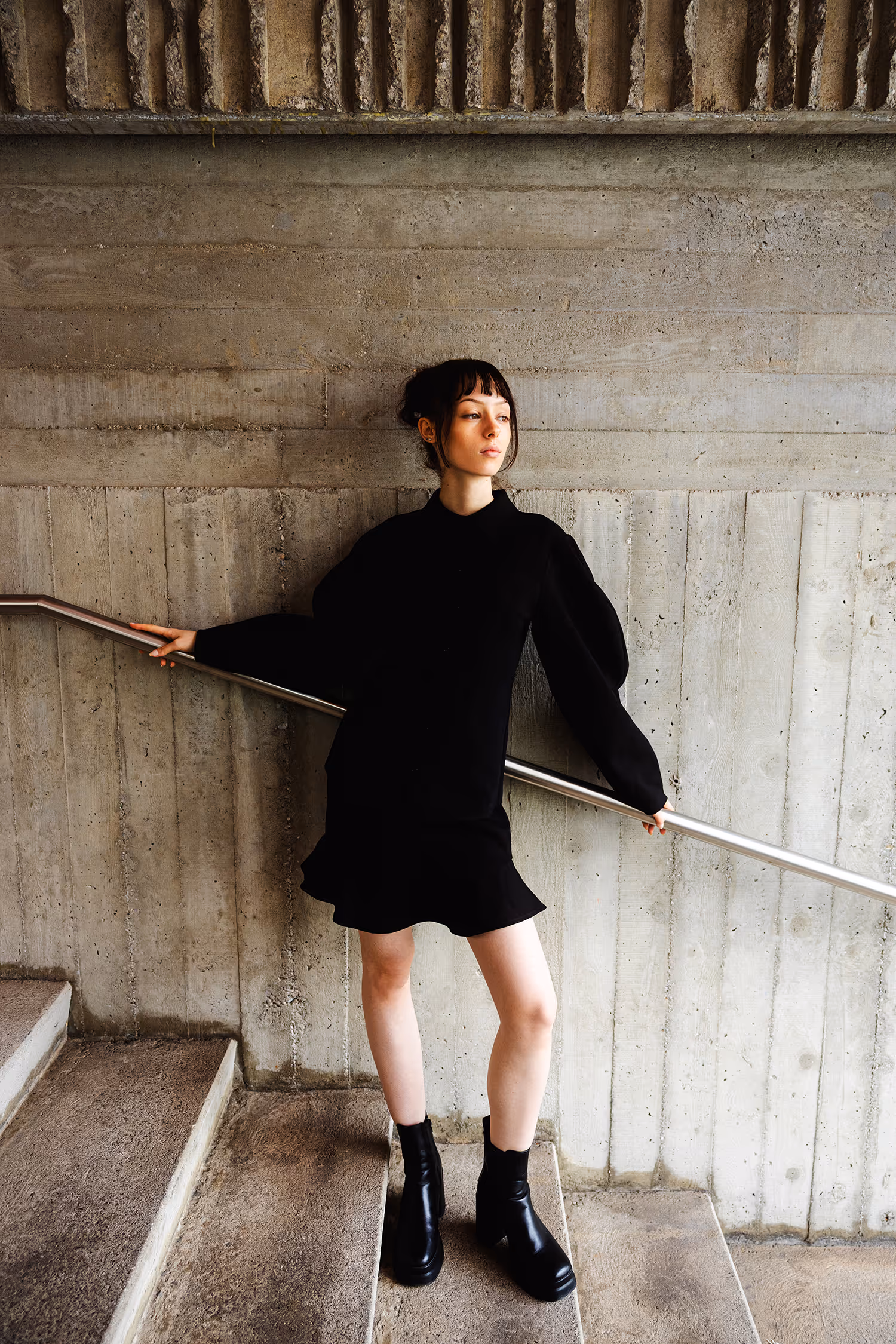 Fashion portrait in Innsbruck featuring brutalist architecture. A model poses in a black minimalist outfit against a raw textured concrete wall and metal railing.