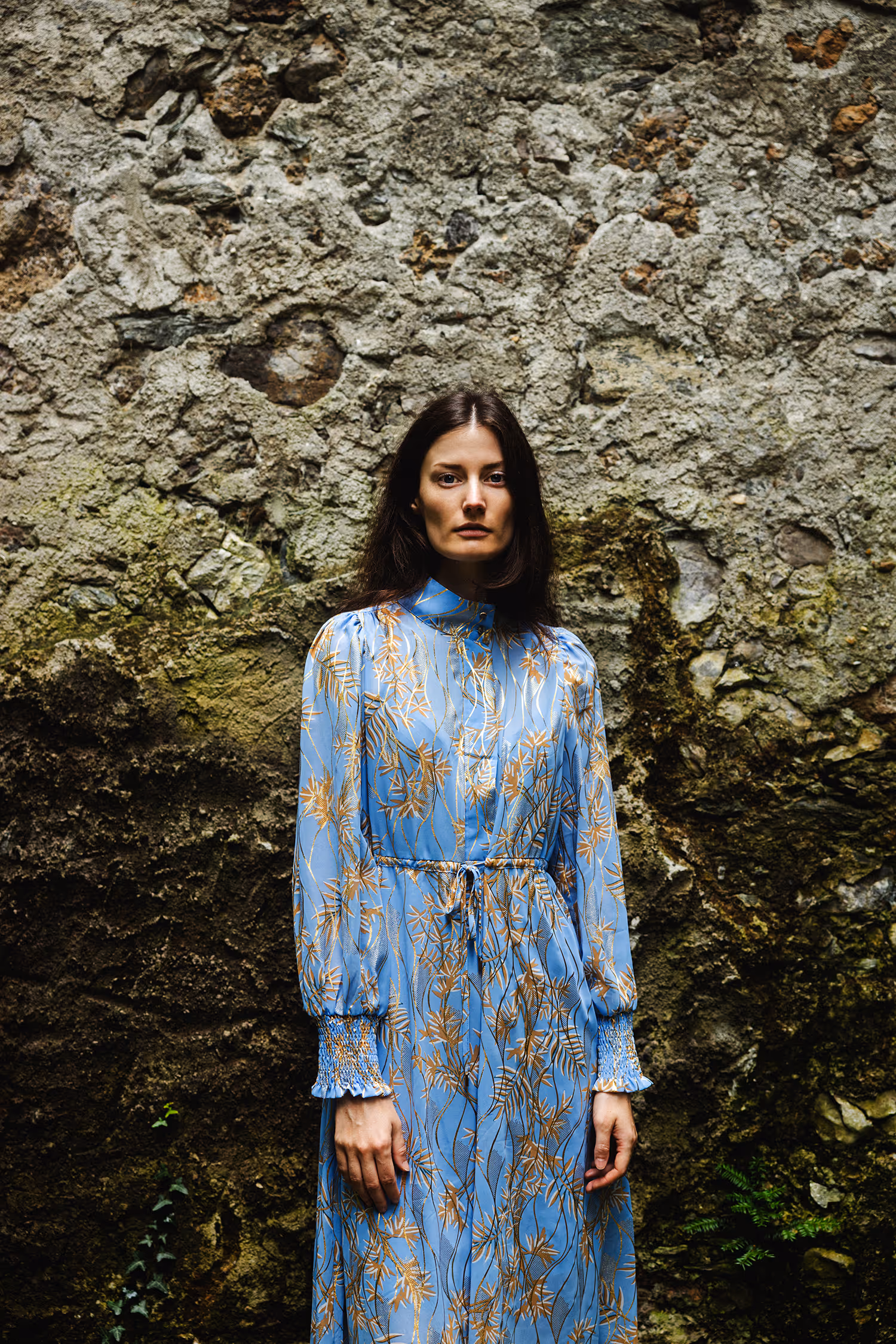Fashion portrait of Viktoria Machajdik at Innsbrucker Hofgarten. She wears a blue botanical dress creating a textural contrast against a rough, mossy stone wall in natural light.