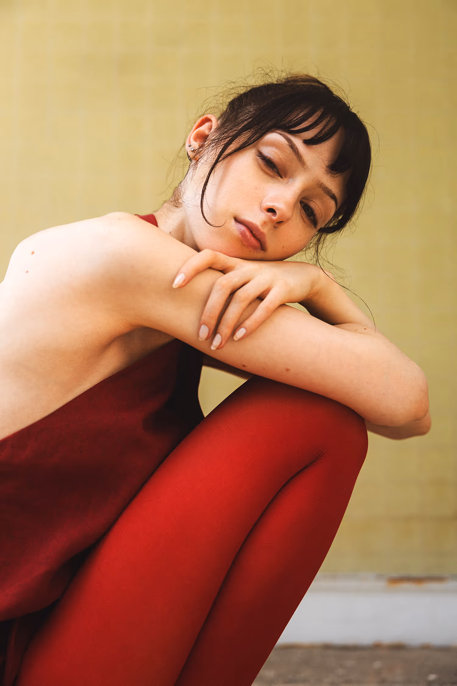 Intimate fashion portrait shot in Innsbruck featuring a model in a deep red monochrome outfit against a yellow wall, showcasing color-blocking and soft natural light.