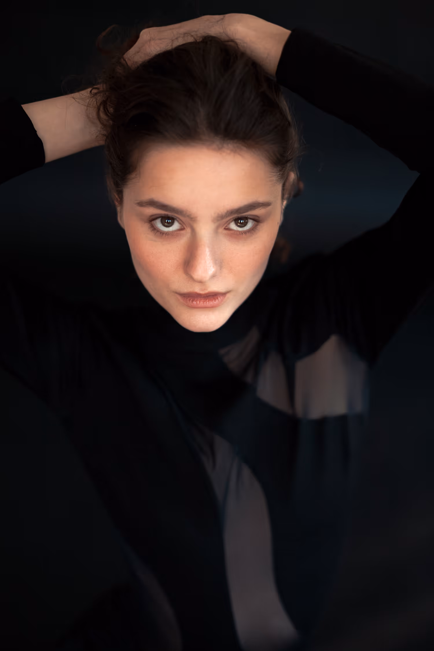 Dramatic low-key fashion portrait featuring a model with an intense direct gaze. She poses with hands behind her head against a dark background, wearing a black outfit with sheer cutout details.