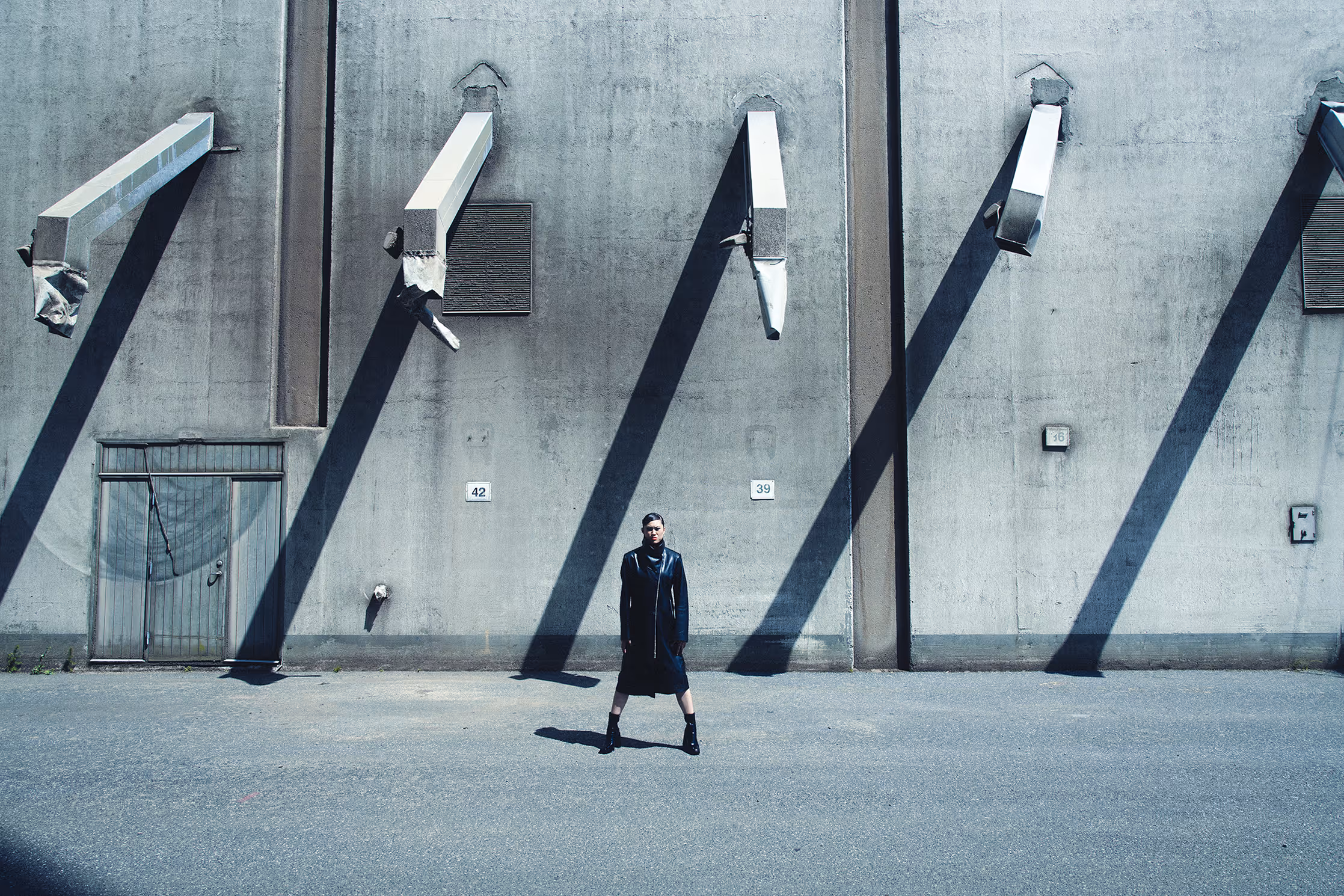 Wide-angle editorial shot set against brutalist industrial architecture in Aarhus. A model in a black leather coat stands amidst dramatic diagonal shadows cast by hard sunlight on a concrete wall.