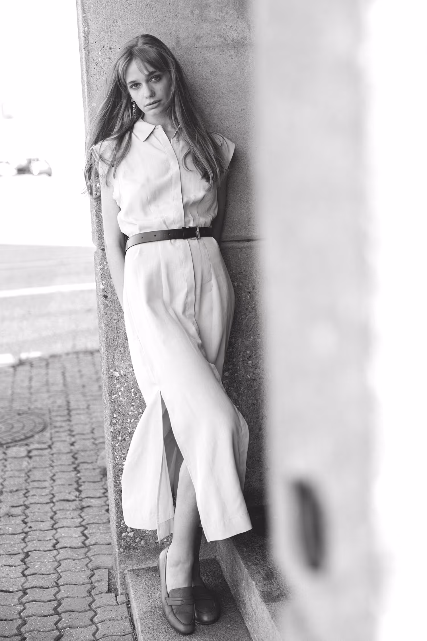 Monochrome street fashion portrait in Aarhus, featuring a model in a minimal white maxi dress and belt, leaning against an urban concrete structure with a soft-focus background.