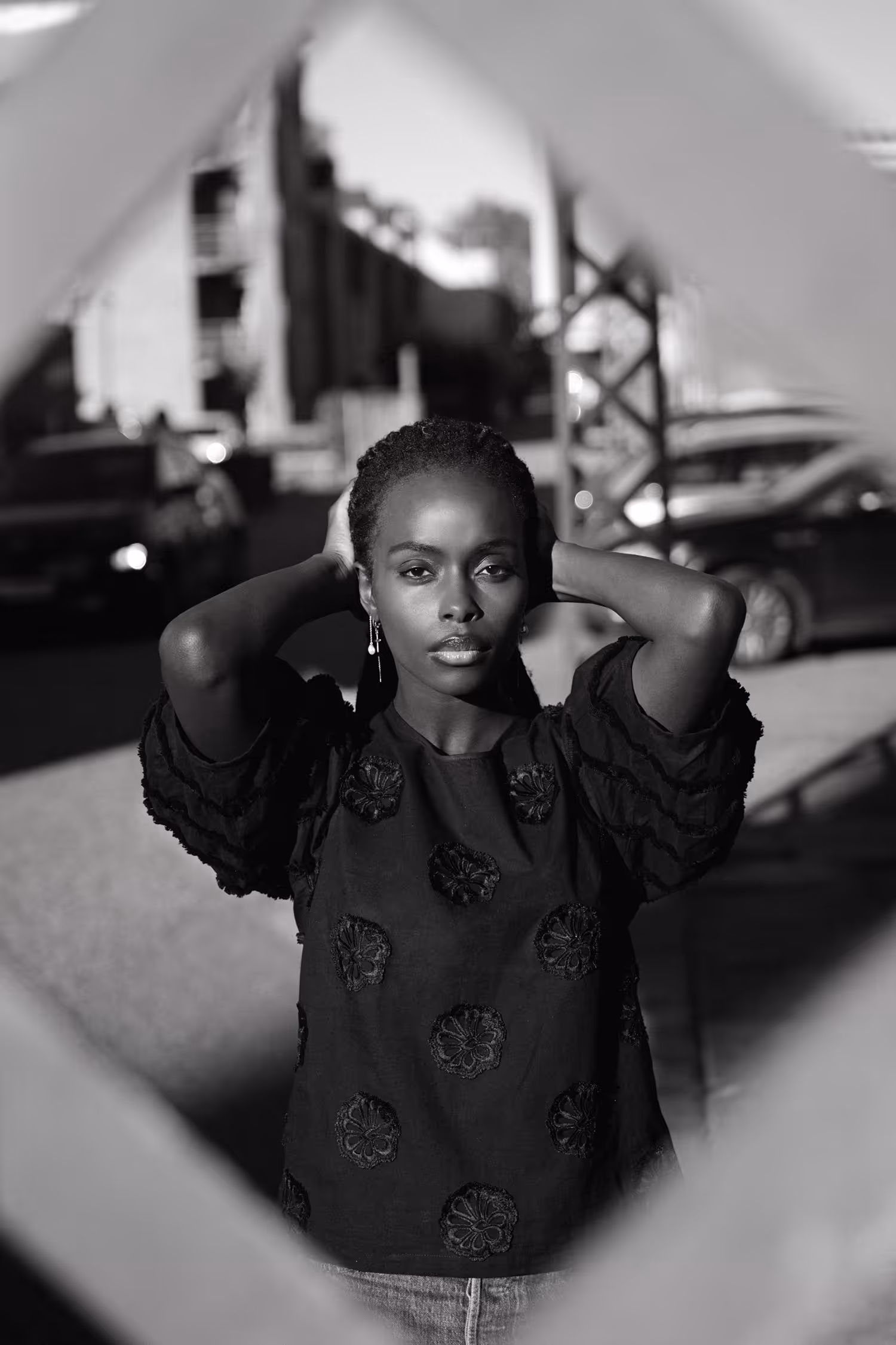 Striking black and white street fashion portrait featuring a model in an embroidered blouse. The image uses hard light and high-contrast, framed by a foreground element, creating a cinematic editorial look.