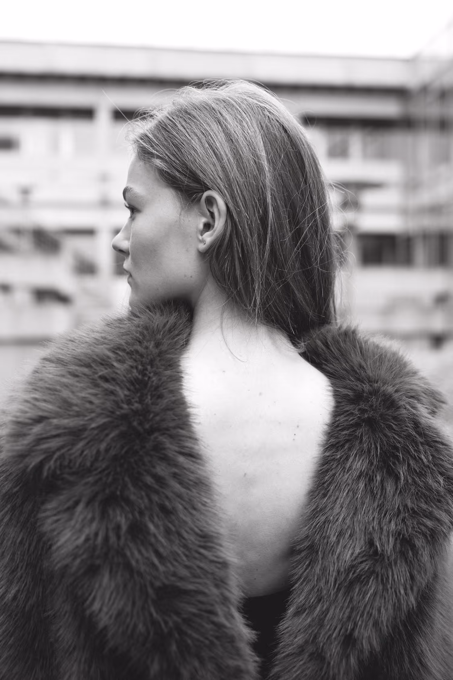 Black and white editorial portrait in Vejle. A model is captured in profile wearing a textured fur coat with an open back, set against a soft-focus architectural background with cinematic grain.
