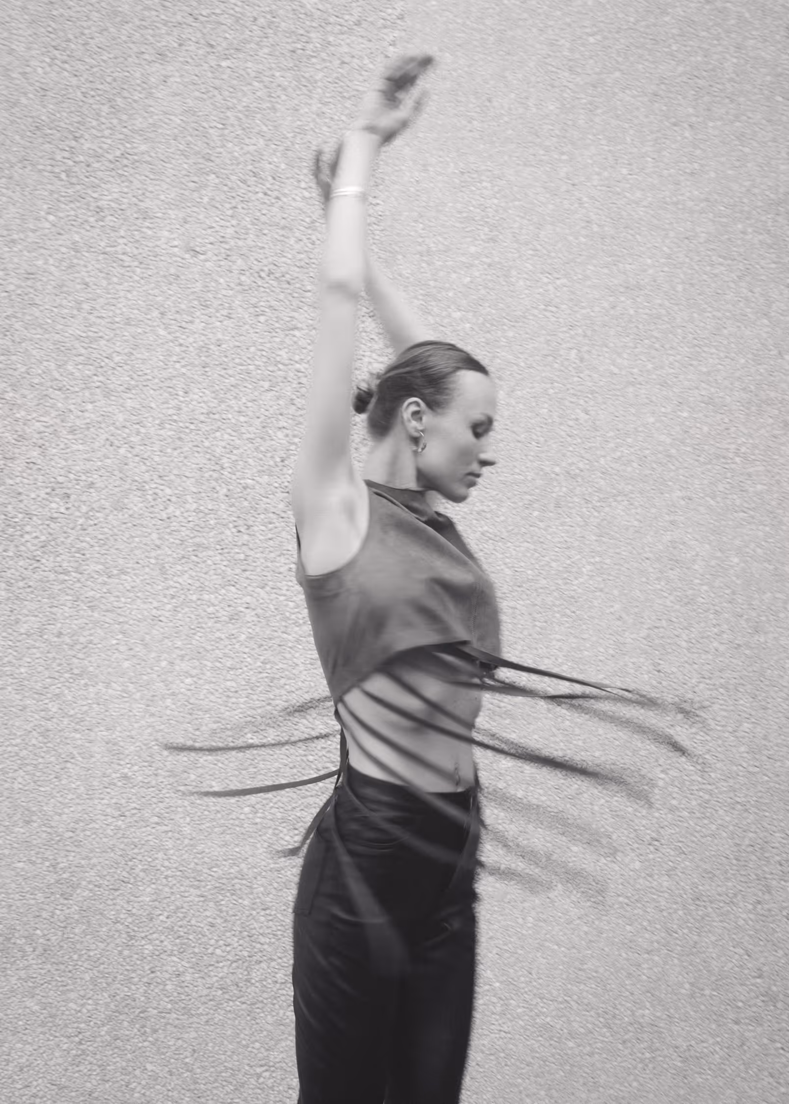 Dynamic B&W fashion editorial shot in Vejle. A model spins with raised arms, creating an artistic motion blur effect with her fringed top against a textured wall.