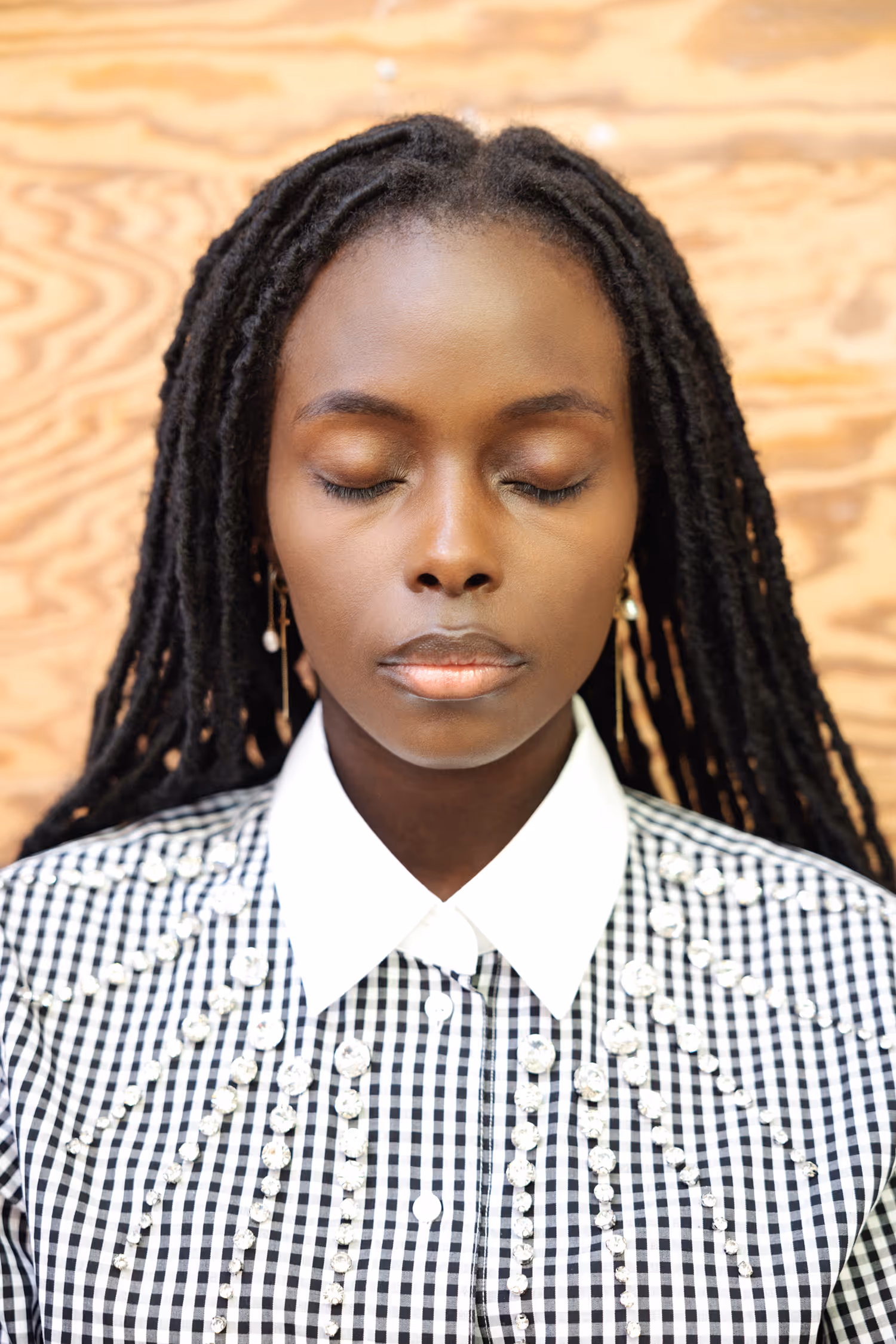 Serene fashion portrait shot in Vejle featuring a Black model with locs and closed eyes, wearing an embellished gingham shirt against a warm wood texture.