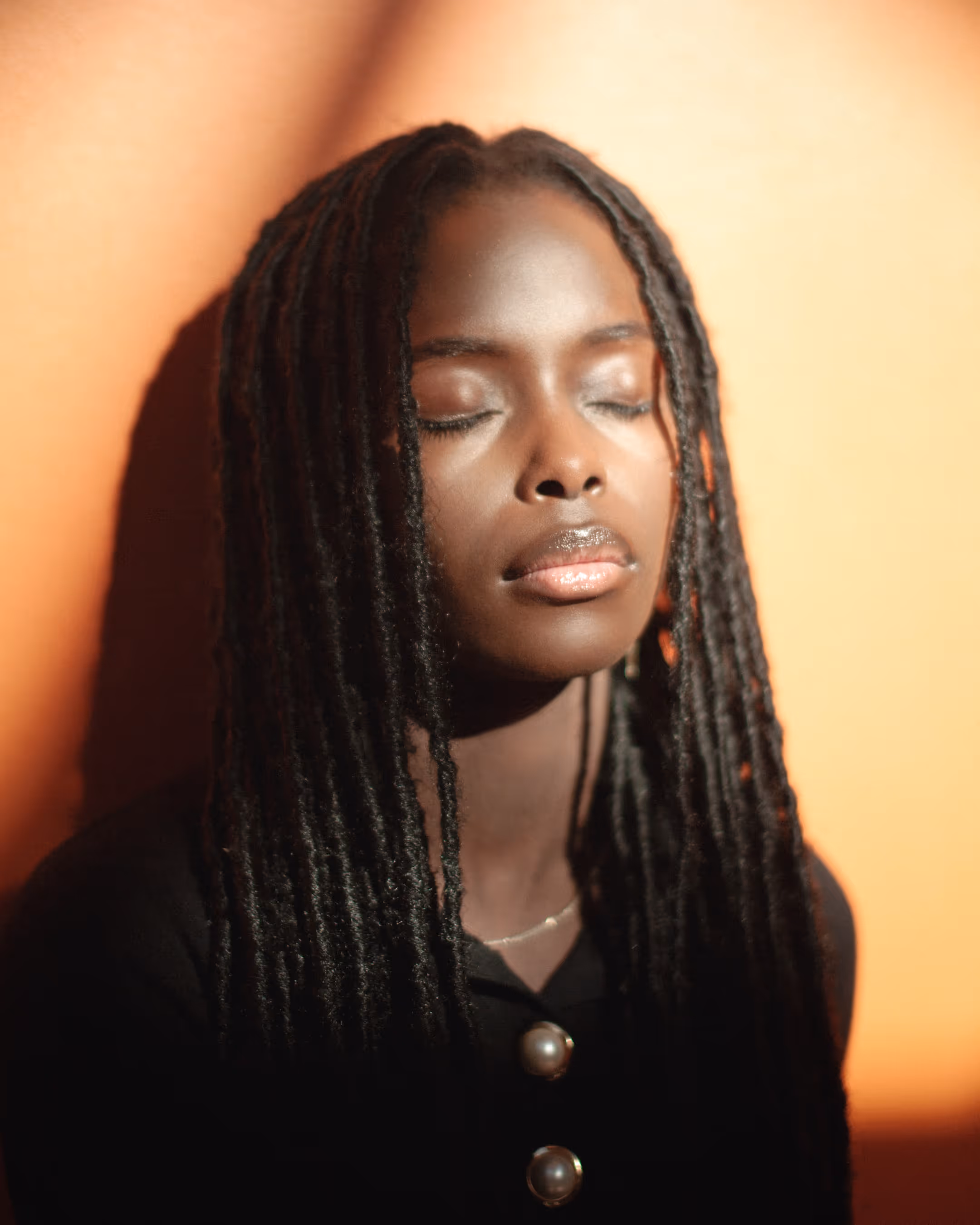 Intimate portrait of a Black model with locs closing her eyes, bathed in warm hard light against an orange wall at Spinderihallerne, Vejle.